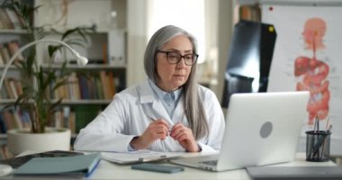 Yaşlı kadın doktor, ofisinde online tıbbi danışmanlık yaparken endişeli görünüyor. Gözlüklü ve beyaz önlüklü olgun bir kadın hastayla iletişim kurarken laptop kullanıyor..