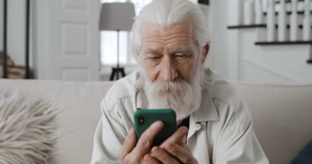 Närbild av gamla gråhåriga man chatta med familjen medan du tittar på telefon sreen.Glada skäggiga manliga pensionär läsa meddelande och leende medans sitter på soffan hemma. — Stockvideo