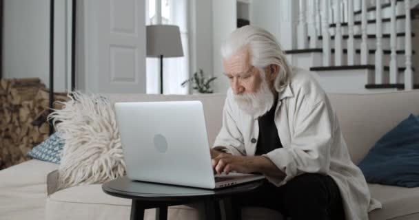 Vista de la cosecha de ancianos de pelo gris abuelo escribiendo mientras se comunica en línea. Hombre barbudo jubilado usando el ordenador portátil mientras está sentado en el sofá en casa. Concepto de ocio y jubilación. — Vídeo de stock