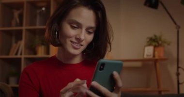 Evdeki kanepede otururken mutlu bir kadının telefon ekranına dokunuşunu ve kayışını yakından izle. Güzel bir kadın sosyal medya bağlantılarını kontrol etmek için akıllı telefon kullanıyor..