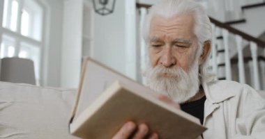 Okumayı bitirirken yaşlı sakallı adamın kapanış kitabını kapat. Yaşlı emeklinin evde koltukta otururken gözlerini ovuşturması ve yorgun görünmesi. Emeklilik kavramı, boş zaman..