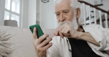 Gri saçlı dedemin haber okurken ekranda gezinip baktığı ekin görüntüsü. Evde koltukta otururken akıllı telefon kullanan yaşlı sakallı adam. Emeklilik kavramı..