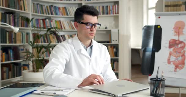 Молодий лікар закриває ноутбук і знімає окуляри, сидячи за столом. Перевантажений чоловік у професійному білому пограбуванні виглядає втомленим і зворушливим, працюючи в медичному кабінеті . — стокове відео