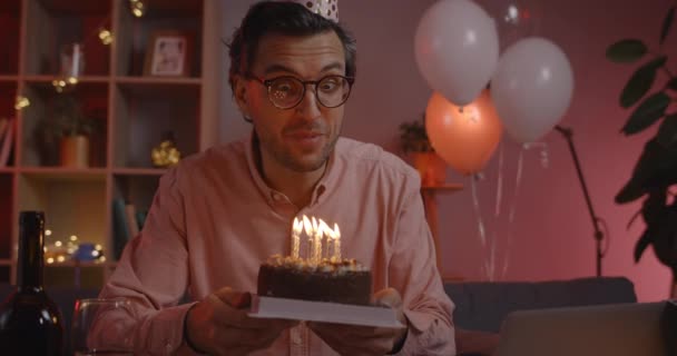 Widok plonu pozytywnej osoby męskiej w kapeluszu urodzinowym śpiew i rozmowy podczas patrzenia na ekranie laptopa. Przystojny mężczyzna w okularach trzyma tort ze świecami siedząc na kanapie. — Wideo stockowe