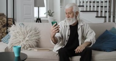 Yaşlı emeklilerin el kol hareketleri, konuşmaları ve gülümsemeleri video görüşmeleri sırasında. Çevrimiçi sohbet ederken akıllı telefon kullanan ve evdeki kanepede oturan neşeli emekliler..