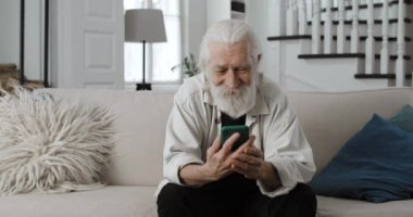 Yaşlı gri saçlı dedemin telefon sapına dokunurken fotoğraflara baktığı mahsul manzarası. Sakallı, mesaj okuyan ve evde koltukta otururken gülümseyen olumlu erkek emekli..