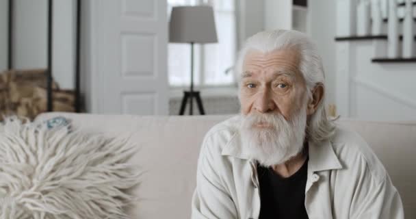 Widok zboża przemyślany stary siwy człowiek patrząc na bok, a nie do kamery. Brodaty emeryt siedzący na kanapie w przytulnym domu. Pojęcie emerytury, ludzi i emocji. — Wideo stockowe
