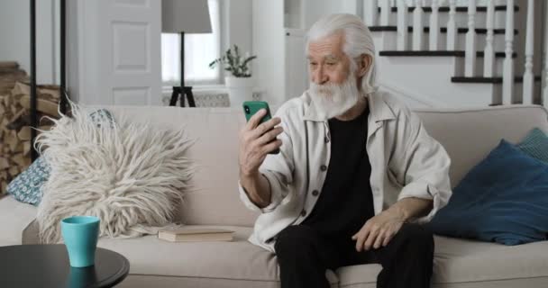 Crop view of senior retiree gesturing, talking and smiling while having video call. Cheerful pensioner using smartphone while having online conversation and sitting on sofa at home. — Stock Video