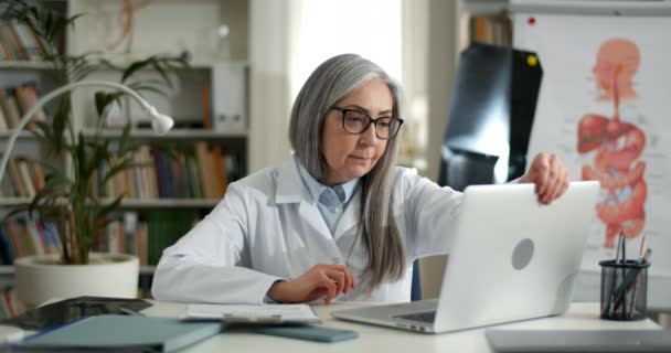 Mulher de óculos e vestido de abrir laptop e começou a digitar ao inserir informações do formulário.Médica idosa trabalhando com computador pessoal e documentos em consultório médico. — Vídeo de Stock