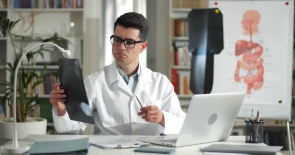 Muž lékař držící rentgen fotografie, zatímco on-line lékařské konzultace v kanceláři. Muž v brýlích a bílý Rob pomocí notebooku při komunikaci s pacientem a sedí u stolu. — Stock video
