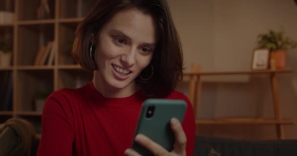 Vista de cerca de la hermosa mujer joven usando el teléfono inteligente y sonriendo mientras está sentado en el sofá en casa. Morena alegre persona femenina desplazándose noticias de alimentación mientras navega por Internet. — Vídeo de stock