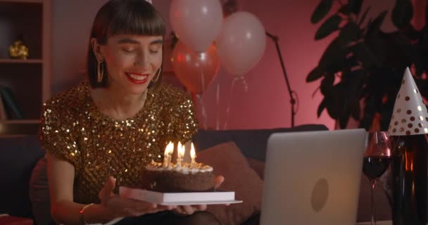 Crop view ofbeautiful woman in birthday hat rejoicing and talking while looking at laptop screen. Positive woman with nose ring blowing candles on cake while sitting at home. — Stock Video