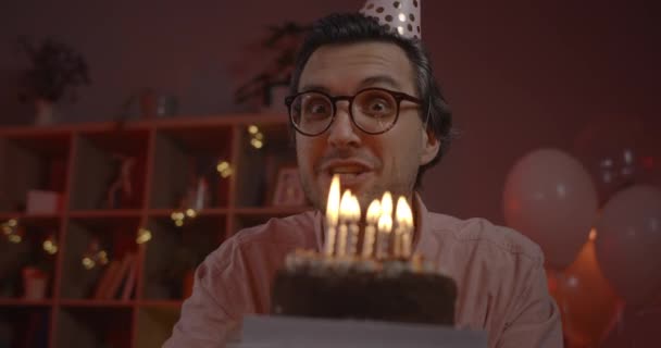 Doğum günü şapkalı heyecanlı adamın kameraya bakarken dilek dilerken görüntüsü. Cheerfu 'lu gözlüklü adam evde otururken konuşuyor ve pasta tutuyor. Çevrimiçi parti kavramı. — Stok video