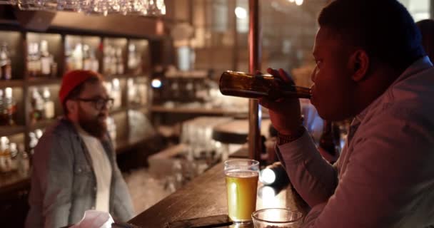 Vista lateral del chico afroamericano bebiendo cerveza de la botella mientras está sentado en el mostrador. Hombre serio pasar tiempo libre y relajarse en el pub. Concepto de ocio. — Vídeo de stock