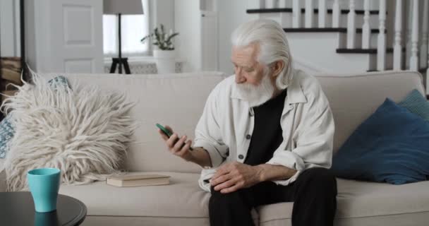 Starszy brodaty mężczyzna mający złe wieści na smartfonie, wyglądający na zdenerwowanego i rozczarowanego. Siwy mężczyzna emeryt odkładając telefon na bok siedząc na kanapie w domu.Pojęcie samotności. — Wideo stockowe