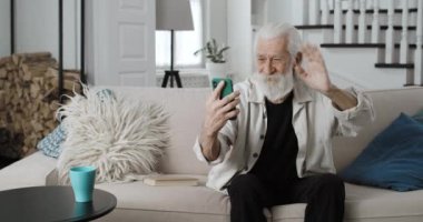 Yaşlı gri saçlı adamın video görüşmesi yaparken el sallaması ve gülümsemesi. Neşeli erkek emekliler evdeki kanepede otururken internet üzerinden iletişim kurmak için akıllı telefon kullanıyorlar..