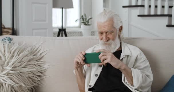 Sentimental male pensioner holding phone horizontally and looking at screen while watching video. Senior bearded man smiling while using smartphone and sitting on couch at home. — Stock Video