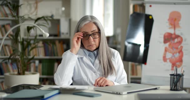 Elderly female doctor thinking and shaking head while sitting at table with laptop on it. Lady in white rob taking off glasses and looking thoughtful while working in medical office. — Stock Video
