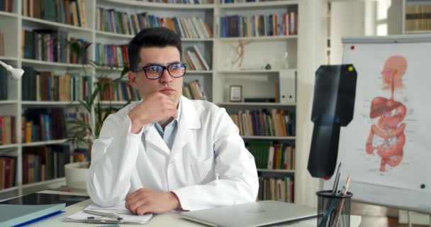 Jovem médico masculino em óculos olhando para o lado e esfregando o queixo enquanto sentado e colocando a mão na mesa. Homem de vestido profissional branco olhando pensativo enquanto trabalhava no escritório médico. — Vídeo de Stock