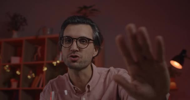Crop view of positive man in glasses holding wineglass and waving bye while having online conversation. Handsome male person talking and looking to camera while sitting at home. — Stock Video