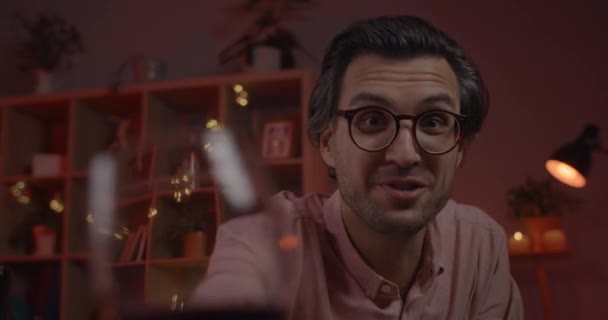 Vrolijke man in bril kijken en juichen voor de camera met wijnglas tijdens het hebben van online gesprek. Knappe man die praat en wijn drinkt terwijl hij 's avonds thuis zit. — Stockvideo