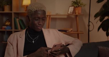 Akıllı telefon kullanan ve evdeki kanepede otururken gülümseyen neşeli Yuoung 'un manzarası. Saçları boyalı Afro-Amerikan erkek haberleri sunuyor ve telefon ekranına bakıyor..