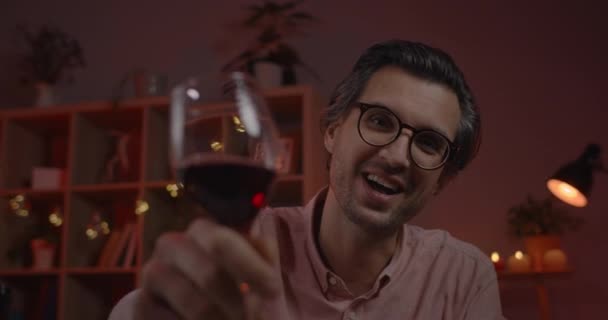 Vista Crop di uomo positivo in bicchieri cercando e tifo per la fotocamera con bicchiere da vino. Bello uomo che parla e si siede a casa mentre fa conversazione online la sera. — Video Stock