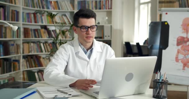 Un joven en blanco roba llenando formulario médico mientras tiene consulta en línea. Guapo médico masculino escuchando al paciente y escribiendo información mientras tiene una conversación en video. — Vídeos de Stock