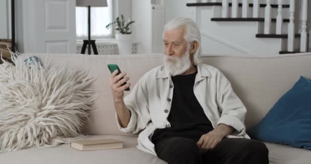 Crop kilátás öreg ősz hajú férfi személy integetett búcsút, és beszél, miközben videohívás. Szakállas nyugdíjas okostelefont használ az online kommunikációhoz, miközben otthon ül a kanapén. — Stock videók