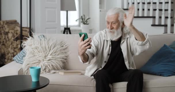 Oříznout pohled na starého šedovlasého muže, jak mává, mluví a usmívá se, zatímco má videohovor. Pozitivní důchodce pomocí smartphone pro on-line komunikaci, zatímco sedí na pohovce doma. — Stock video