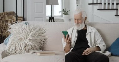 Sakallı emekli erkek, telefonu dikey tutuyor ve video izlerken ekrana bakıyor. Akıllı telefon kullanırken gülümseyen ve evdeki kanepede oturan yaşlı gri saçlı adam..