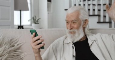 Yaşlı gri saçlı adamın görüntüsü. El sallıyor, konuşuyor ve gülümsüyor. Neşeli erkek emekliler evdeki kanepede otururken internet üzerinden iletişim kurmak için akıllı telefon kullanıyorlar..