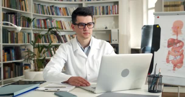 Молодой человек в очках и белый грабитель показывает знак ОК во время онлайн-консультации. Мужчина врач закрывает ноутбук после окончания работы, сидя за столом в медицинском кабинете. — стоковое видео