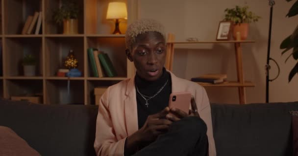 Närbild av manliga afro amerikan person rulla telefonskärm och säger wow när du sitter på soffan hemma. Millennial man ser förvånad och ler när du använder smartphone. — Stockvideo