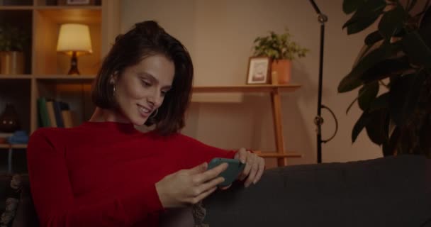 Beskär syn på positiva tusenåriga kvinna tittar på video och skaka huvudet när du sitter på soffan hemma. Attraktiv ung kvinna som håller smartphone horisontellt och tittar på skärmen. — Stockvideo