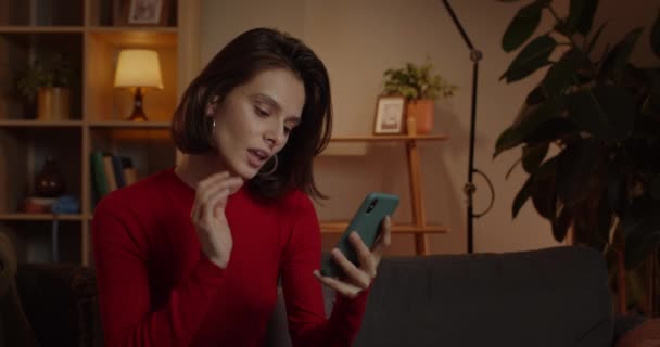 Crop view van verleidelijke vrouw met behulp van smartphone en glimlachen tijdens het zitten op de bank thuis. Brunette millennial vrouwelijke persoon scrollen nieuwsfeed en kijken naar het scherm van de telefoon. — Stockvideo