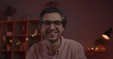 Gözlüklü, yakışıklı, pozitif bir adamın kameraya bakarken el sallaması ve konuşması. Neşeli adam evde otururken internette gülüp sohbet ediyor. İletişim kavramı.