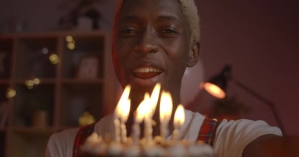 Primo piano di uomo afro americano sorridente in cappello di compleanno gioire e parlare mentre si guarda alla fotocamera. Giovane ragazzo positivo con i capelli tinti soffiando candele sulla torta mentre seduto a casa. — Video Stock