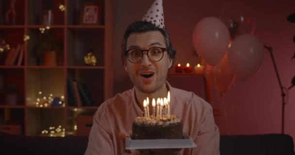 Wesoły młody człowiek w kapeluszu urodzinowym radujący się i śpiewający piosenkę patrząc w kamerę. Przystojny mężczyzna po trzydziestce dmucha świeczki na tort i świętuje siedząc w domu. — Wideo stockowe