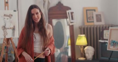 Lviv, Ukrayna - 17 Ocak 2020: Kameraya bakarken başını kaldırıp gülümseyen kadın. Yaratıcı sanat atölyesinde dururken elinde fırça tutan ve poz veren çekici kız ressamın portresi.