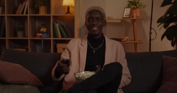Beskär utsikt över millennial glad man tar fjärrkontroll och slå på tv. afro amerikan man person som håller skål med popcorn medan du sitter bekvämt på soffan och titta på film. — Stockvideo