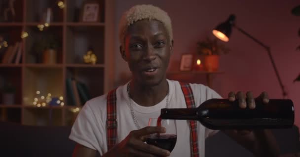 Snygg ung man hälla och dricka vin från vinäger och skratta medan du har online-konversation. Man afro amerikan person pratar och ler medan du tittar på kameran hemma. — Stockvideo