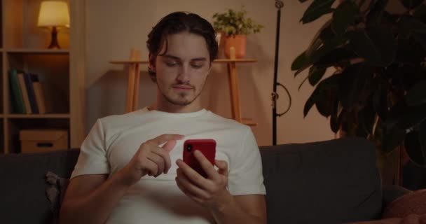 夕方に自宅のソファに座っている間、若い男のクロップビューは、スマートフォンの画面をスクロールします。余暇を楽しみながら携帯電話を利用する白人男性. — ストック動画