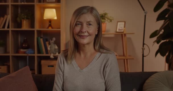 Menutup pandangan wanita dewasa yang bahagia melihat ke kamera. Portrait of joyful female person smile while sitting on sofa at home in evening. Konsep dari orang-orang headshot dan emosi. — Stok Video