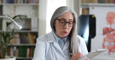 Gözlüklü kadın doktorun video konsültasyonu sırasında elinde tuttuğu ve hastanın tıbbi formunu doldurduğu ekin görüntüsü. Kadın konuşuyor, yazıyor ve tıp bürosunda kameraya bakıyor..