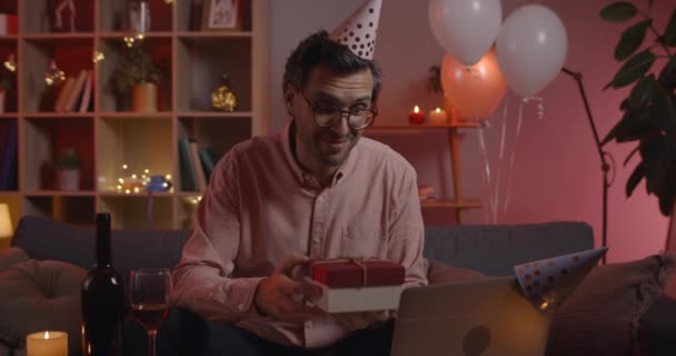 Widok na faceta w okularach mówiącego i siedzącego na kanapie podczas świętowania urodzin online. Pozytywny mężczyzna w papierowej czapce trzyma pudełko prezentów i uśmiecha się podczas rozmowy wideo. — Wideo stockowe
