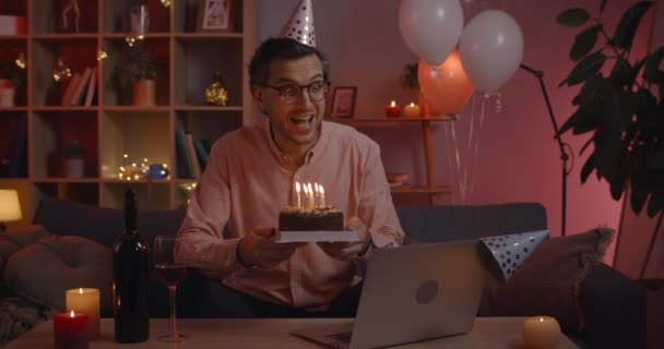 Przystojny mężczyzna w okularach w kapeluszu urodzinowym radujący się i mówiący patrząc na ekran laptopa. Pozytywny mężczyzna trzyma tort ze świecami siedząc na kanapie w domu. — Wideo stockowe