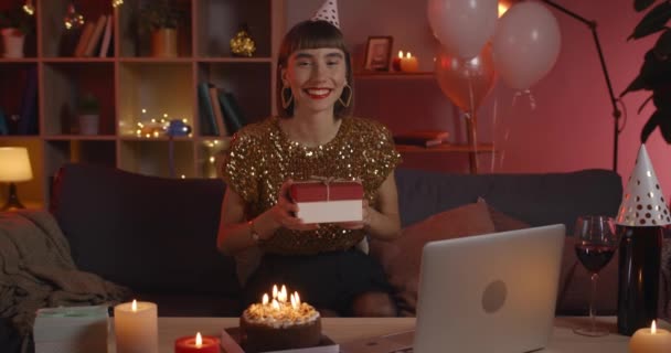 Crop view of attractive female person in birthday hat looking to camera and laughing. Positive millennial woman in stylish clothes holding present box while sitting on couch. — Stock Video