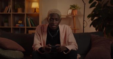Kanepede otururken elinde joystick tutan ve kullanan Afro-Amerikalı genç bir adamın görüntüsü. Milenyum erkeği eğlenirken oyun oynuyor. Boş zaman ve eğlence anlayışı.