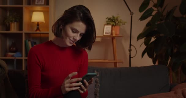 Beskär bild av attraktiv ung kvinna med hjälp av smartphone och leende när du sitter på soffan hemma. Brunett tusenårig kvinnlig person rulla nyhetsflöde samtidigt kontrollera sociala medier. — Stockvideo